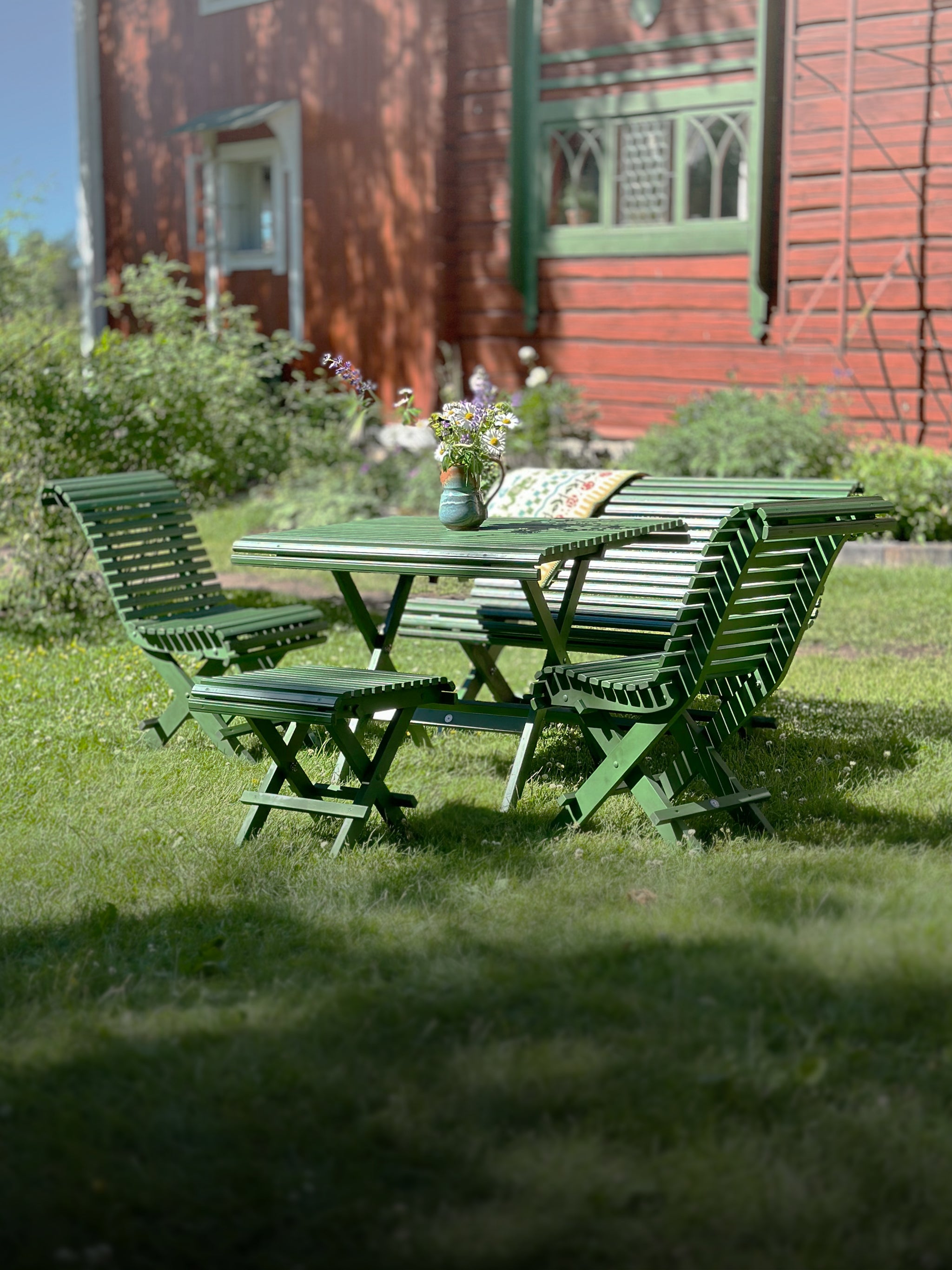 gröna hopfällbara trädgårdsmöbler i Carl Larsson anda – Sundborn grupp med soffa, stol, pall och bord placerat på en gräsmatta framför en röd villa.
