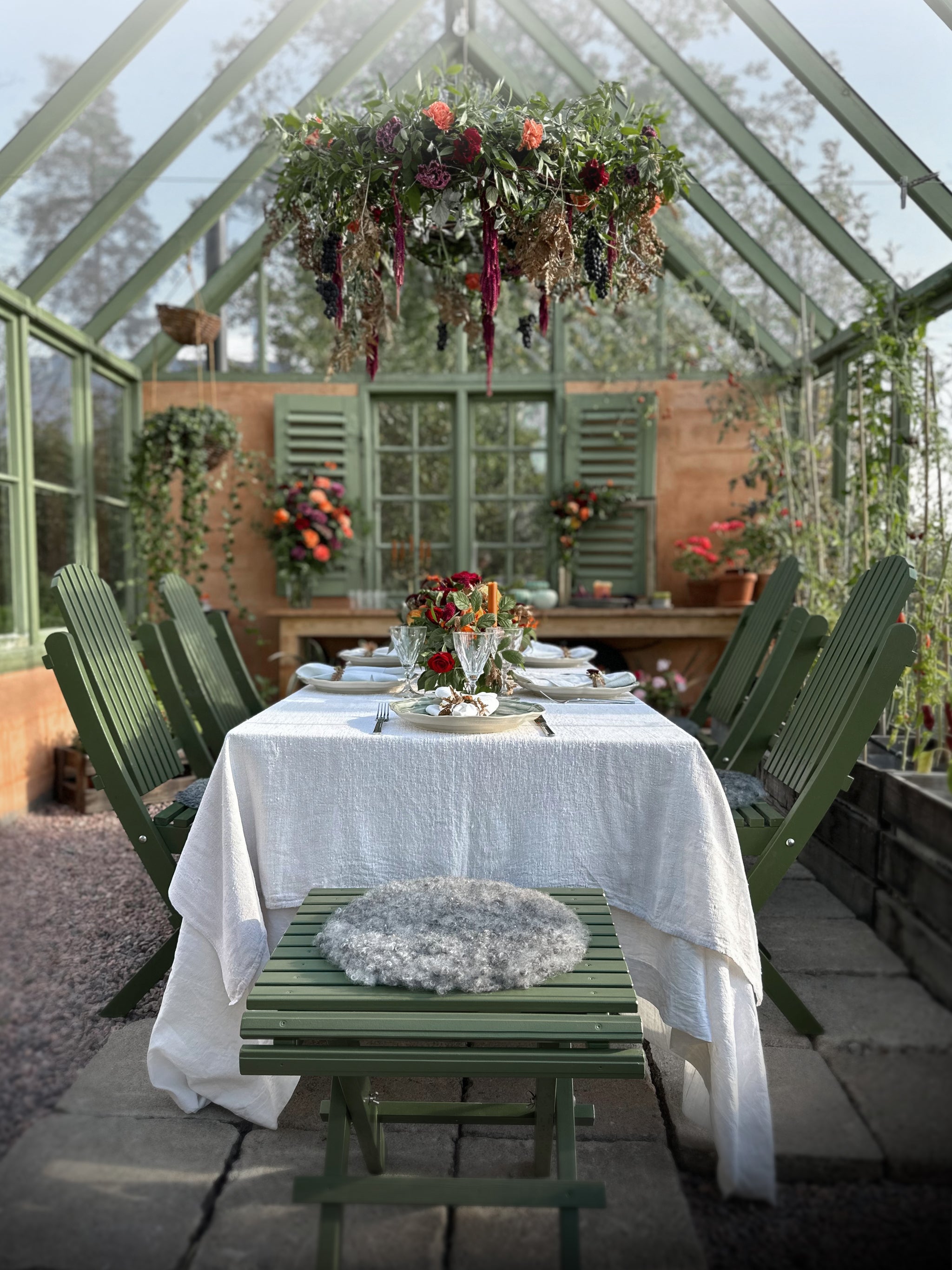 Gröna klassiska trädgårdsmöbler i sekelskiftesstil – hopfällbart matbord och stolar inne i ett växthus, vackert dekorerat med blommor och linneduk.